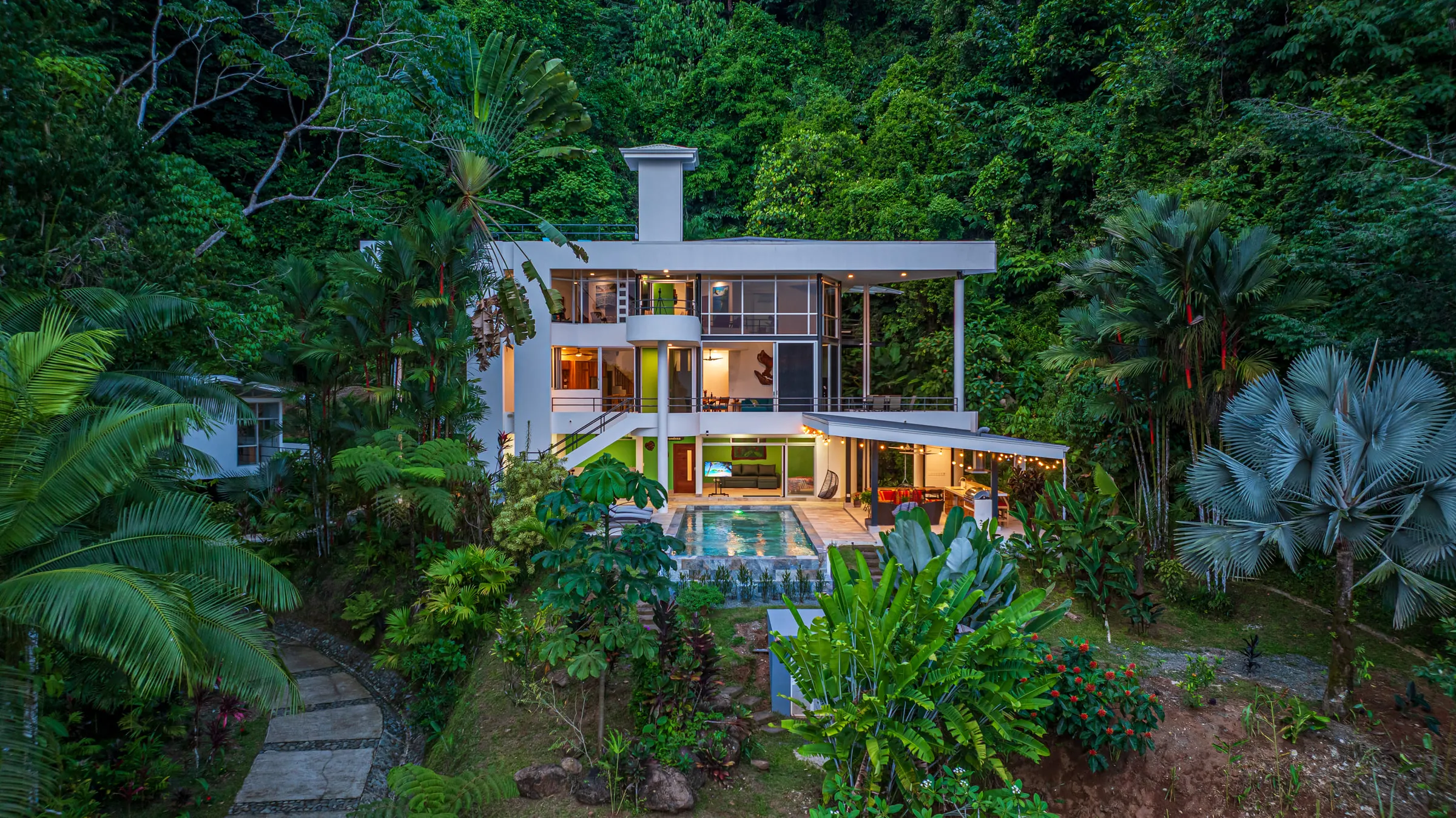 The whole house at dusk, glowing from inside the rainforest, with the pool lit below and the jungle rising behind it